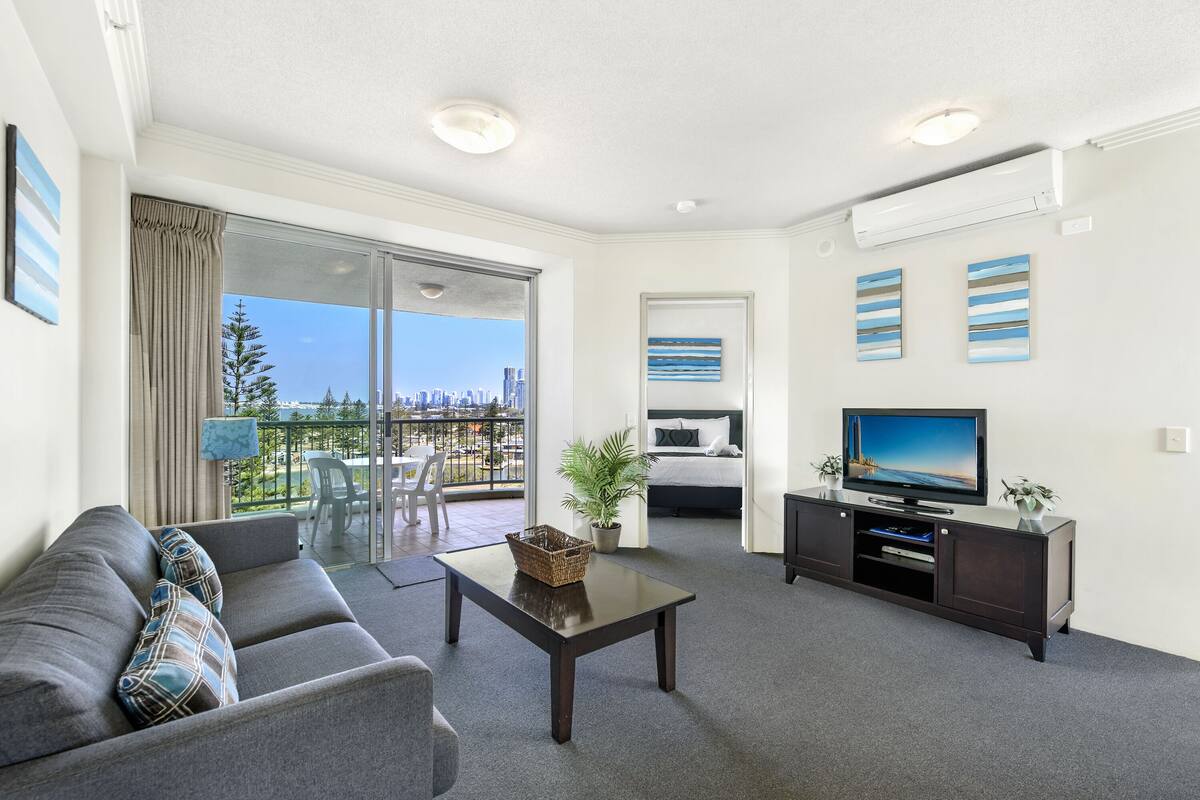 A modern living room with a sofa, a coffee table, a TV on a cabinet, and a balcony with a city view.