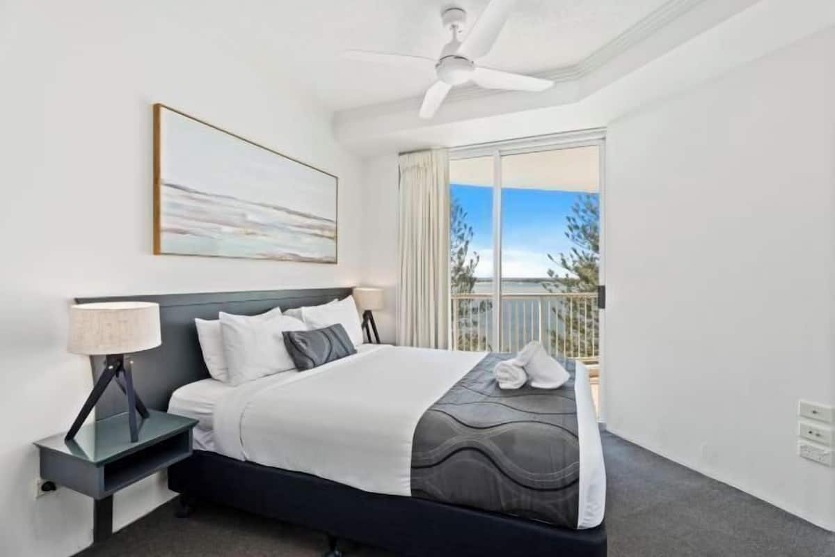 A hotel room with a bed, a nightstand, a ceiling fan, a window with a view, and a painting on the wall.