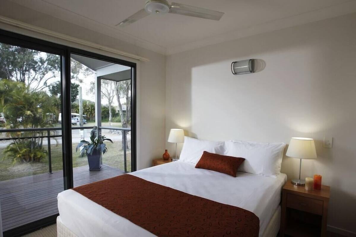 A hotel room with a bed, two bedside lamps, a ceiling fan, and a view of the outdoors.