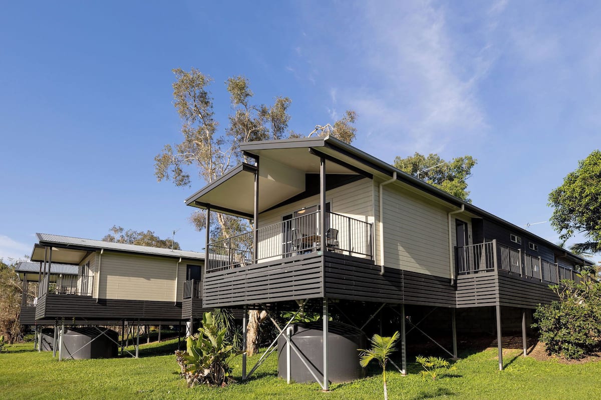 A two-story house with a balcony, elevated on stilts, surrounded by greenery.