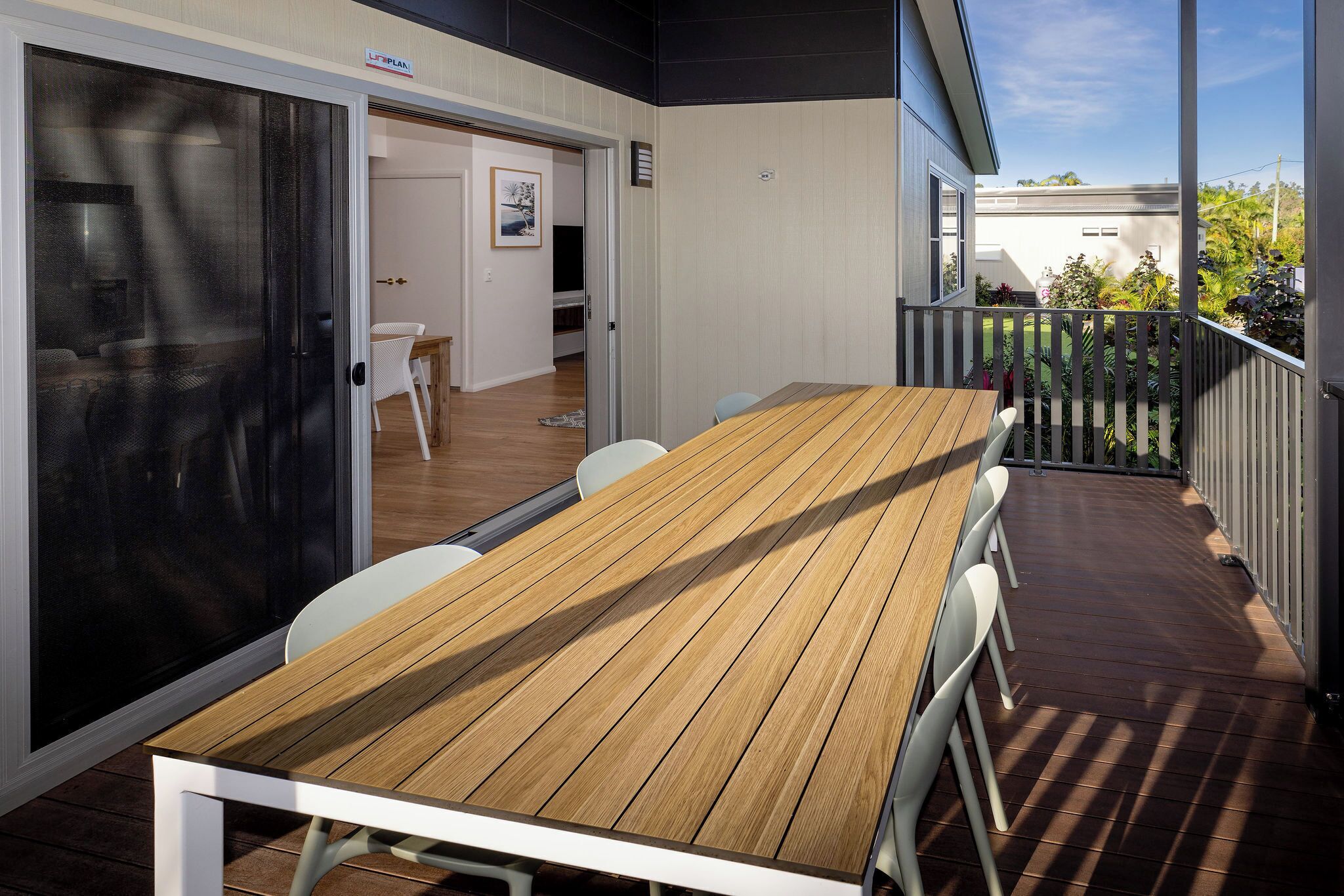 Una terraza de madera con mesa y sillas, un ventanal corredero y vista a una casa y jardín.