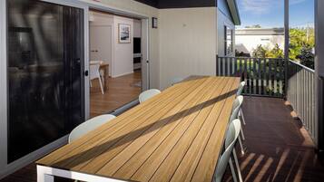 Una terraza de madera con mesa y sillas, un ventanal corredero y vista a una casa y jardín.