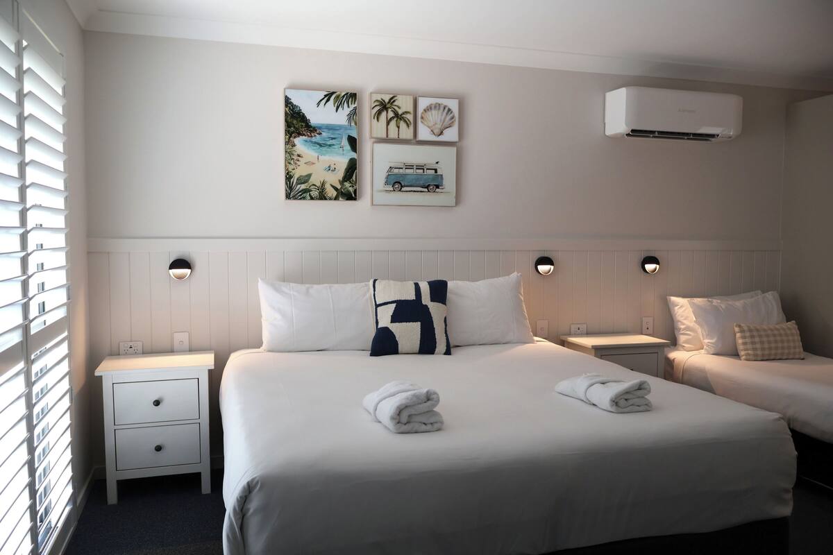 A hotel room with a large bed, two bedside tables, a wall-mounted air conditioner, and artwork depicting a beach scene.