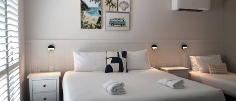 A hotel room with a large bed, two bedside tables, a wall-mounted air conditioner, and artwork depicting a beach scene.