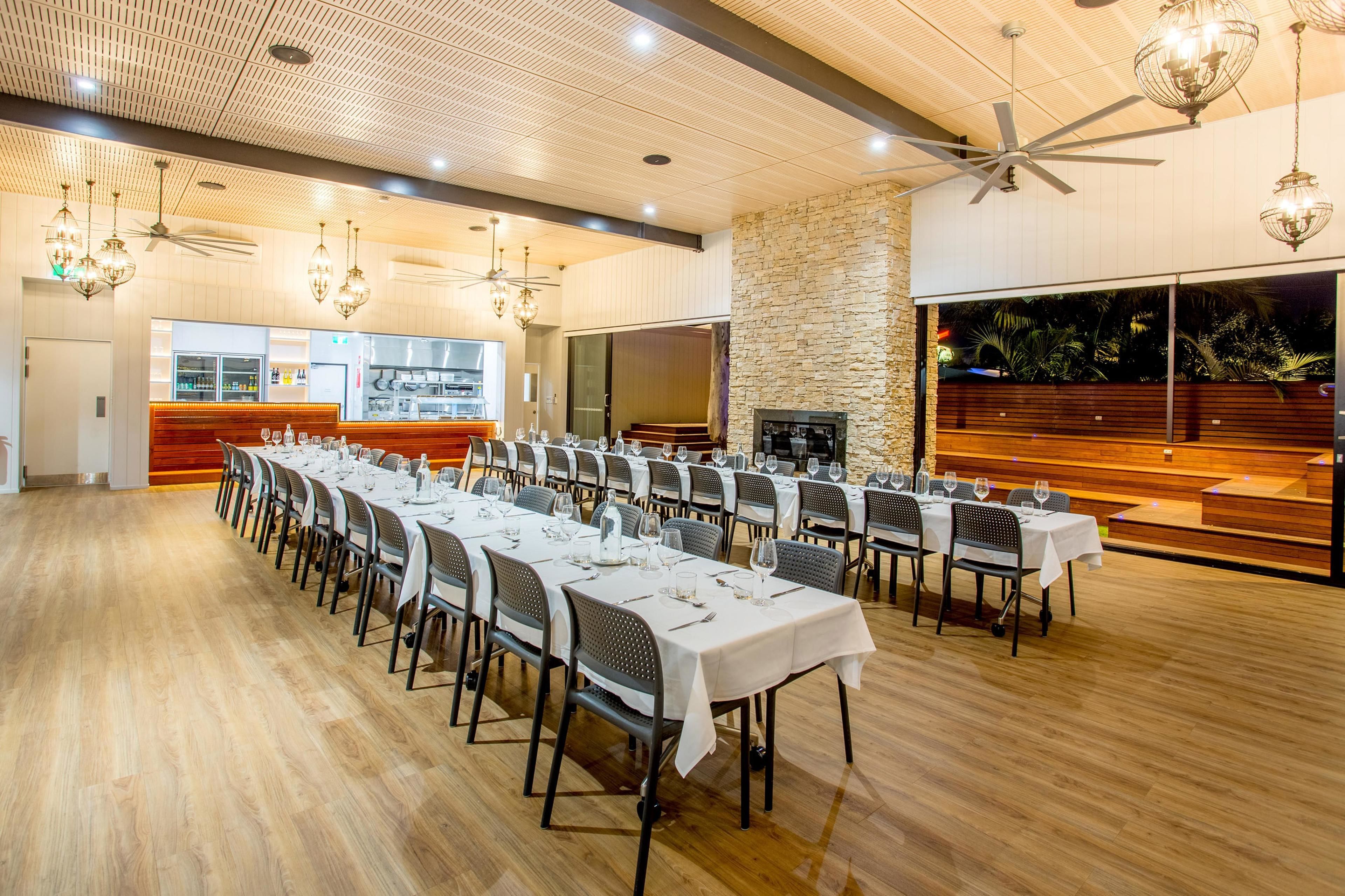A spacious banquet hall with long tables set for dining, a stone accent wall, and a large window overlooking a garden.