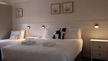 Habitación de hotel con dos camas, ropa de cama blanca, cabecera, luces fijadas a la pared, mesita de noche y cajón.