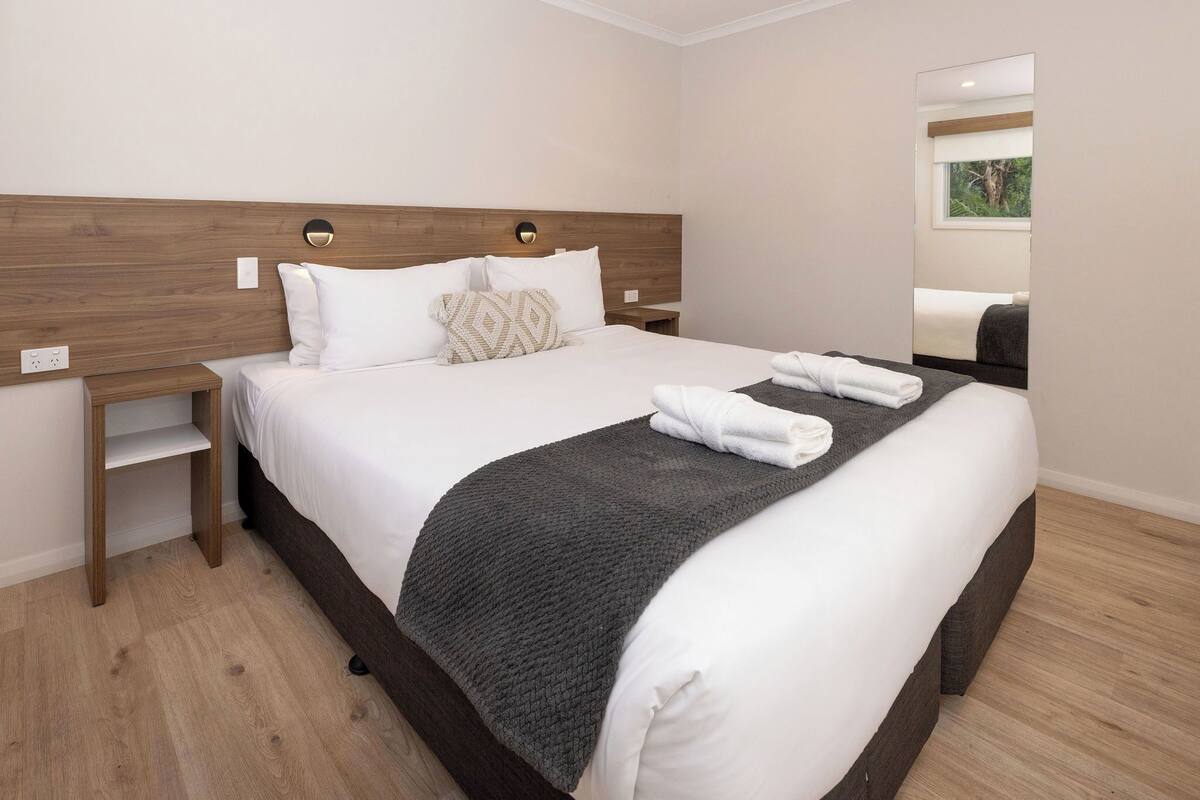 A neatly made bed with white linens and a dark gray blanket, a wooden headboard, bedside tables, and a mirror on the wall.
