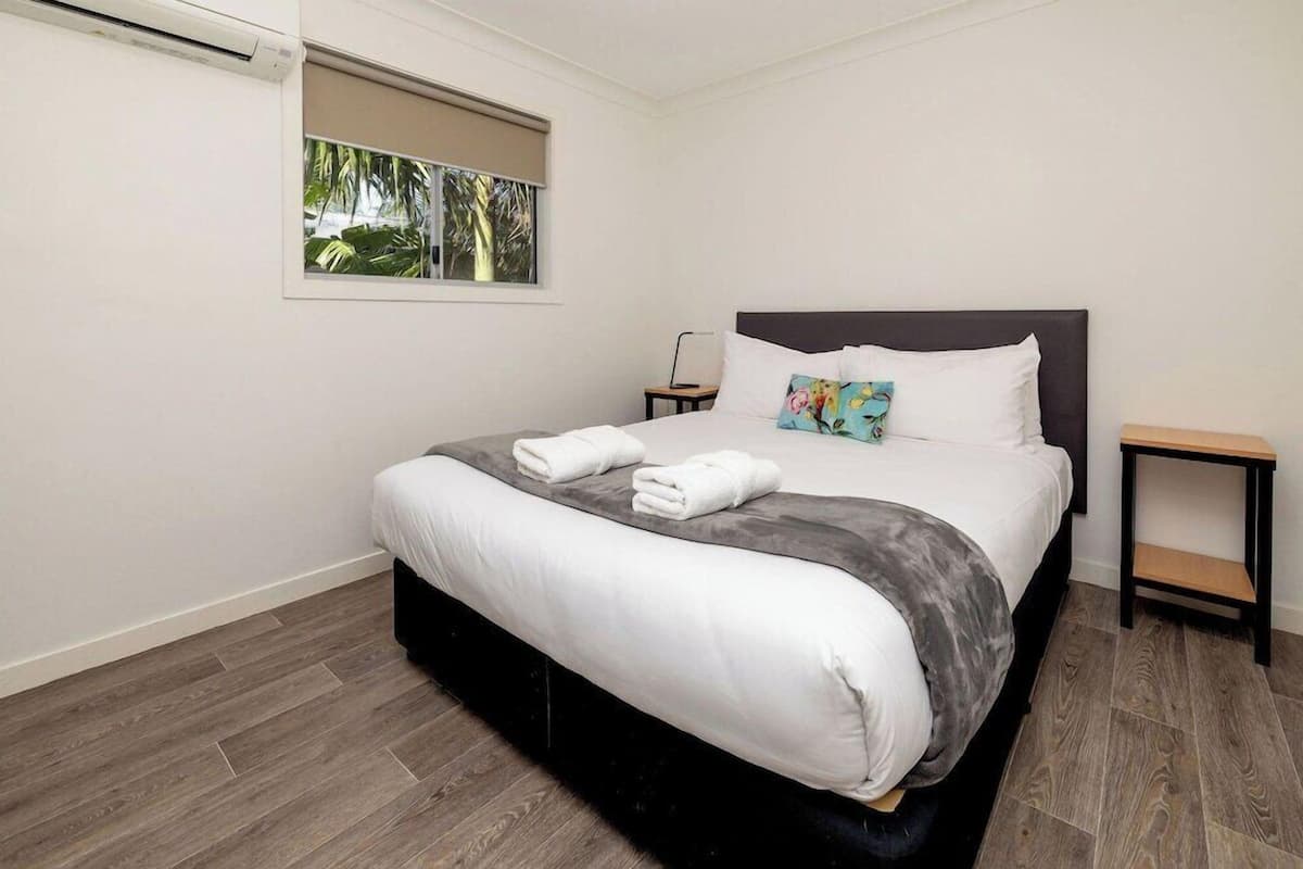 A neatly arranged bedroom with a bed, a bedside table, a nightstand, and a window with a view of greenery.