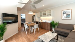 A modern living room with a flat-screen TV, a ceiling fan, a dining area with a wooden table and chairs, a sofa, and a kitchen area with cabinets and a sink.