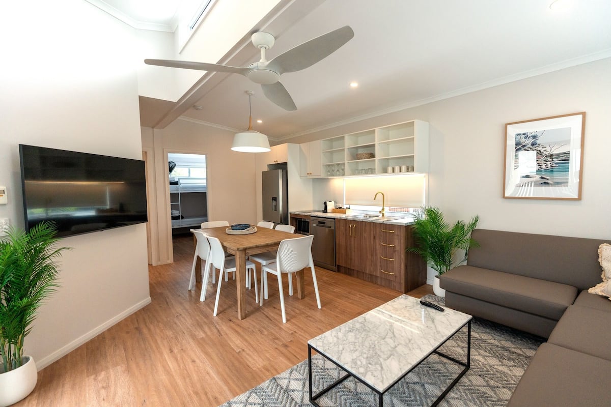 A modern living room with a flat-screen TV, a ceiling fan, a dining area with a wooden table and chairs, a sofa, and a kitchen area with cabinets and a sink.