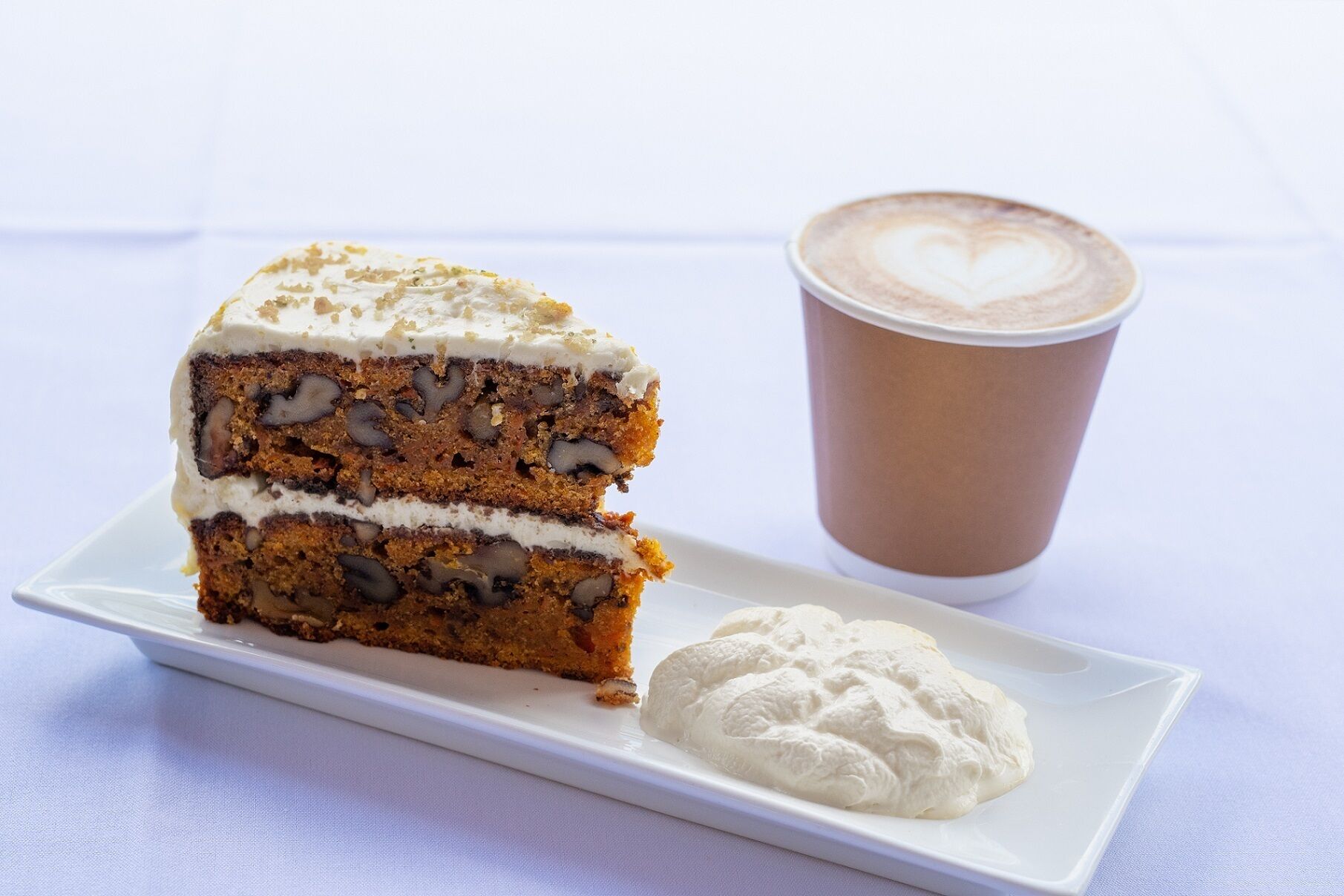 Ein Stück Karottenkuchen mit Sahnefrosting und einer Kugel Schlagsahne auf einem weißen Teller, neben einer Tasse Kaffee mit Latte-Art.