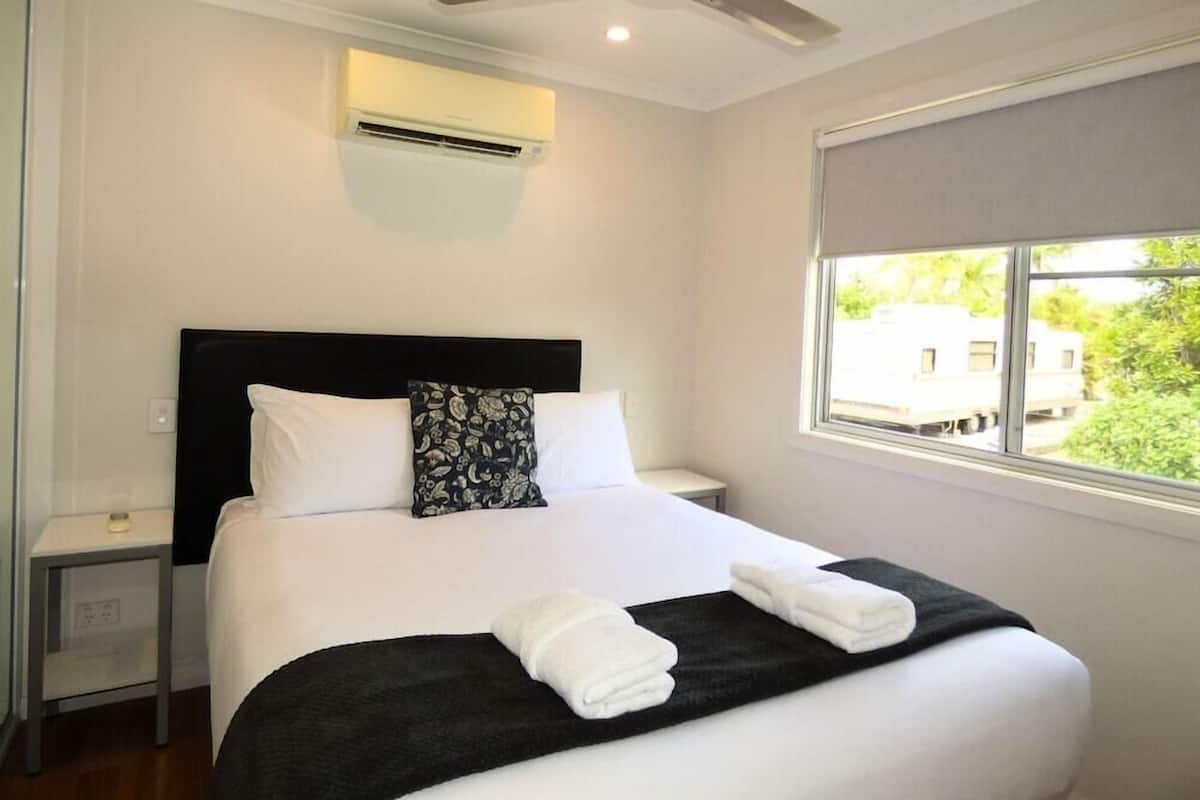 A hotel room with a bed, a ceiling fan, an air conditioning unit, a bedside table, and a window with a view of greenery and a vehicle outside.