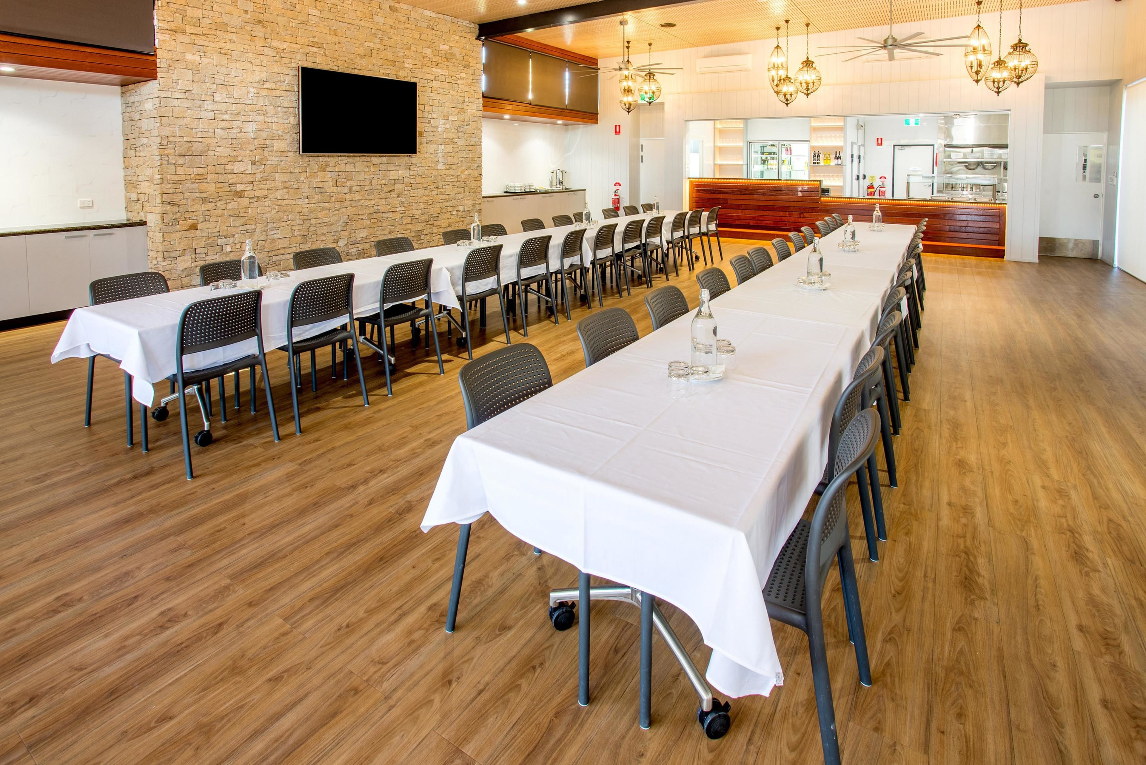 A conference room with a long table, chairs, a projector screen, and a kitchen area in the background.