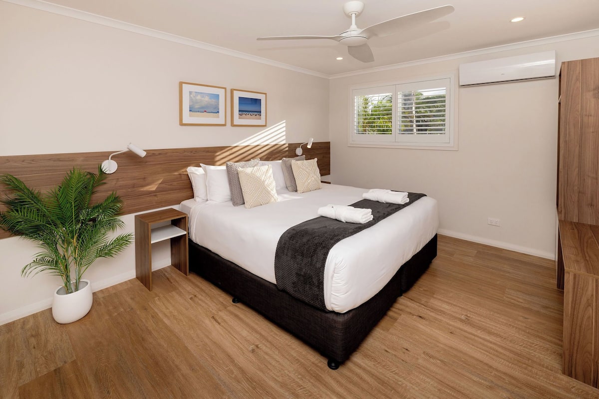 A bedroom with a bed, bedside table, lamp, air conditioning unit, window, and a potted plant.