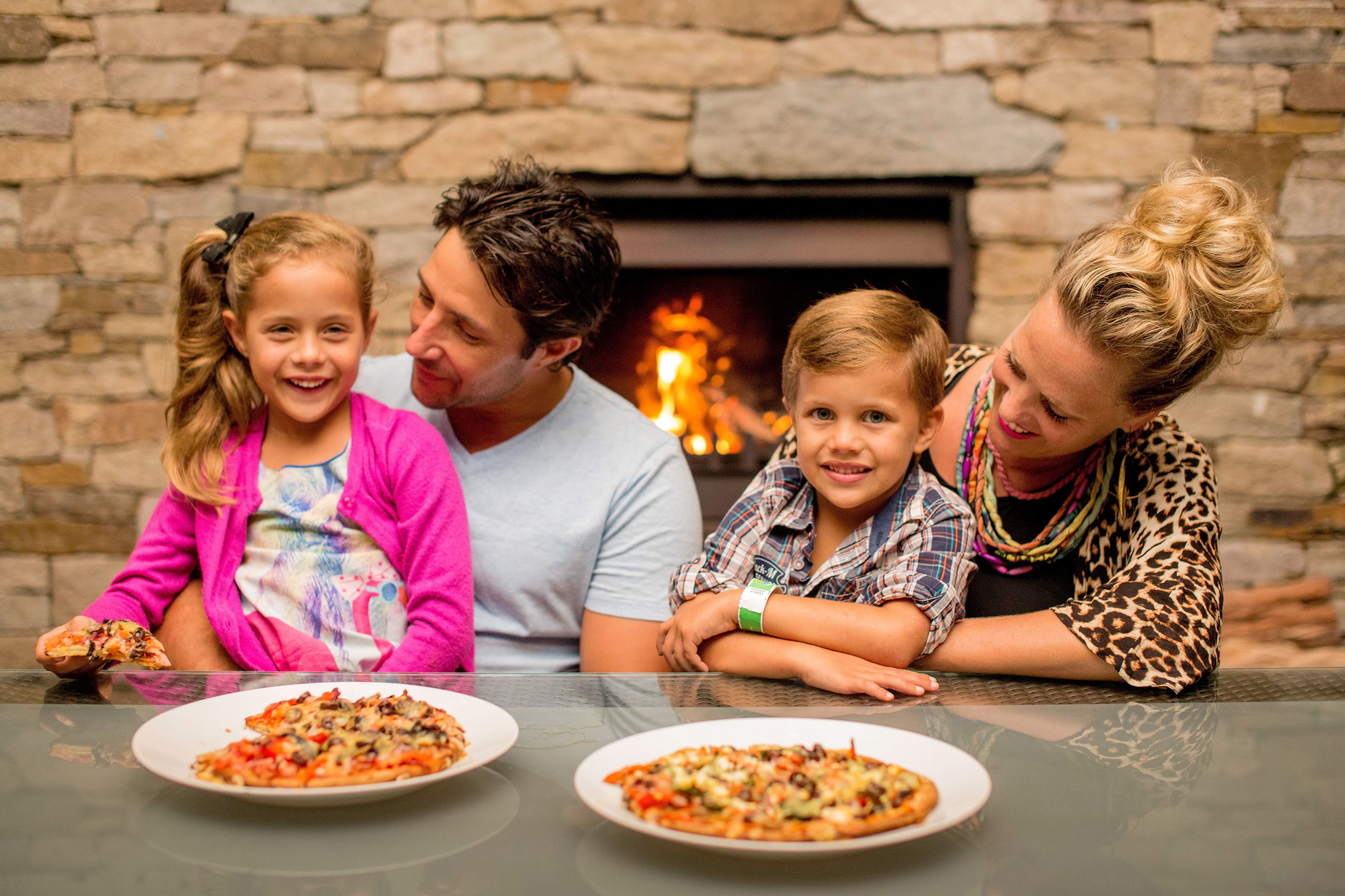 Eine Familie sitzt an einem Tisch vor einem Kamin aus Stein, auf dem Pizza steht.