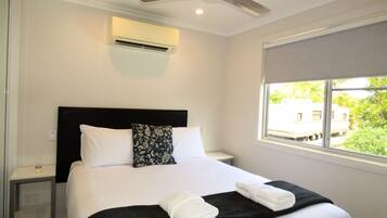 A hotel room with a bed, a ceiling fan, an air conditioning unit, a bedside table, and a window with a view of greenery.