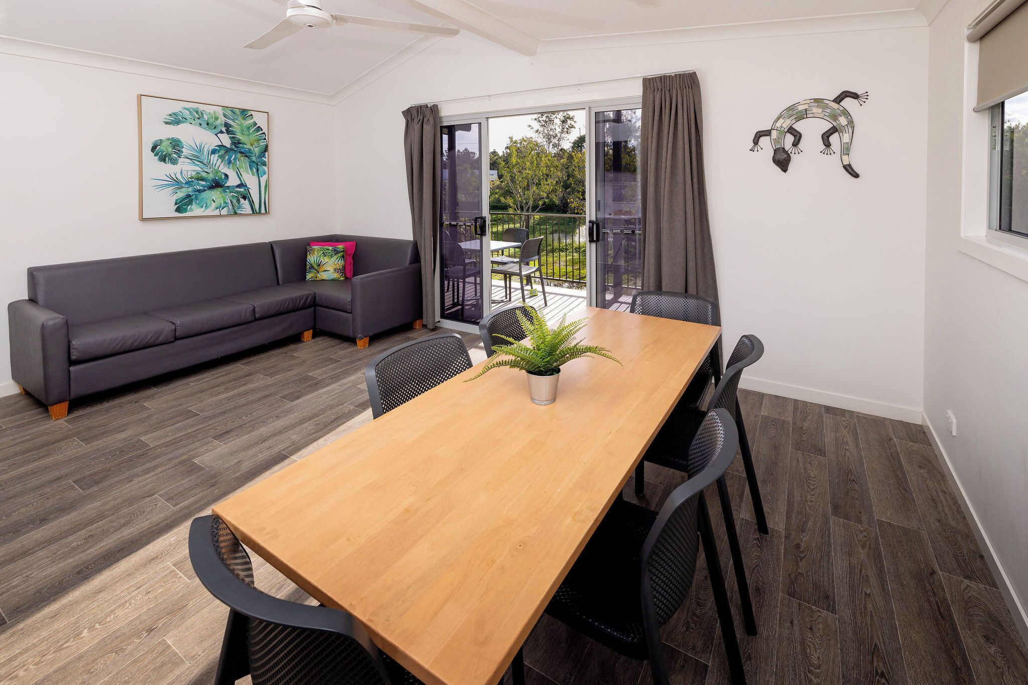 A modern living room with a wooden dining table, black chairs, a gray sofa, and a large window with a view of the outdoors.