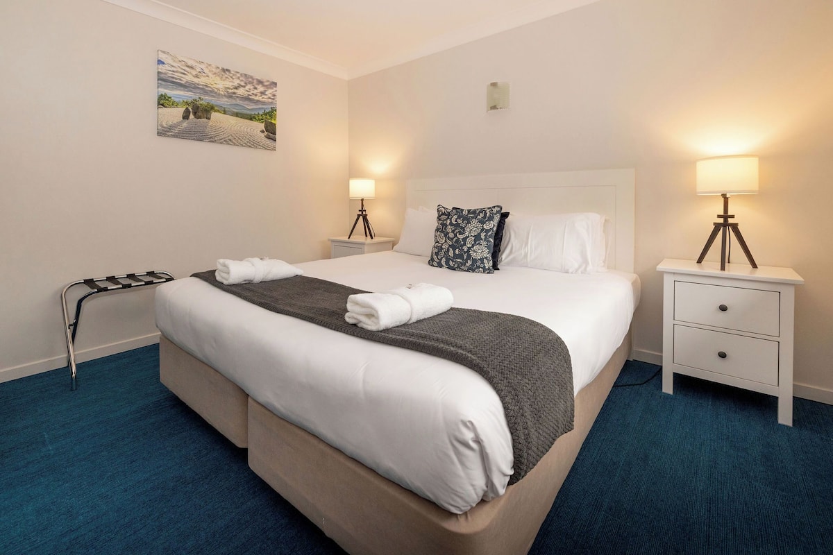 A hotel room with a large bed, two bedside tables with lamps, a painting on the wall, and a blue carpet.