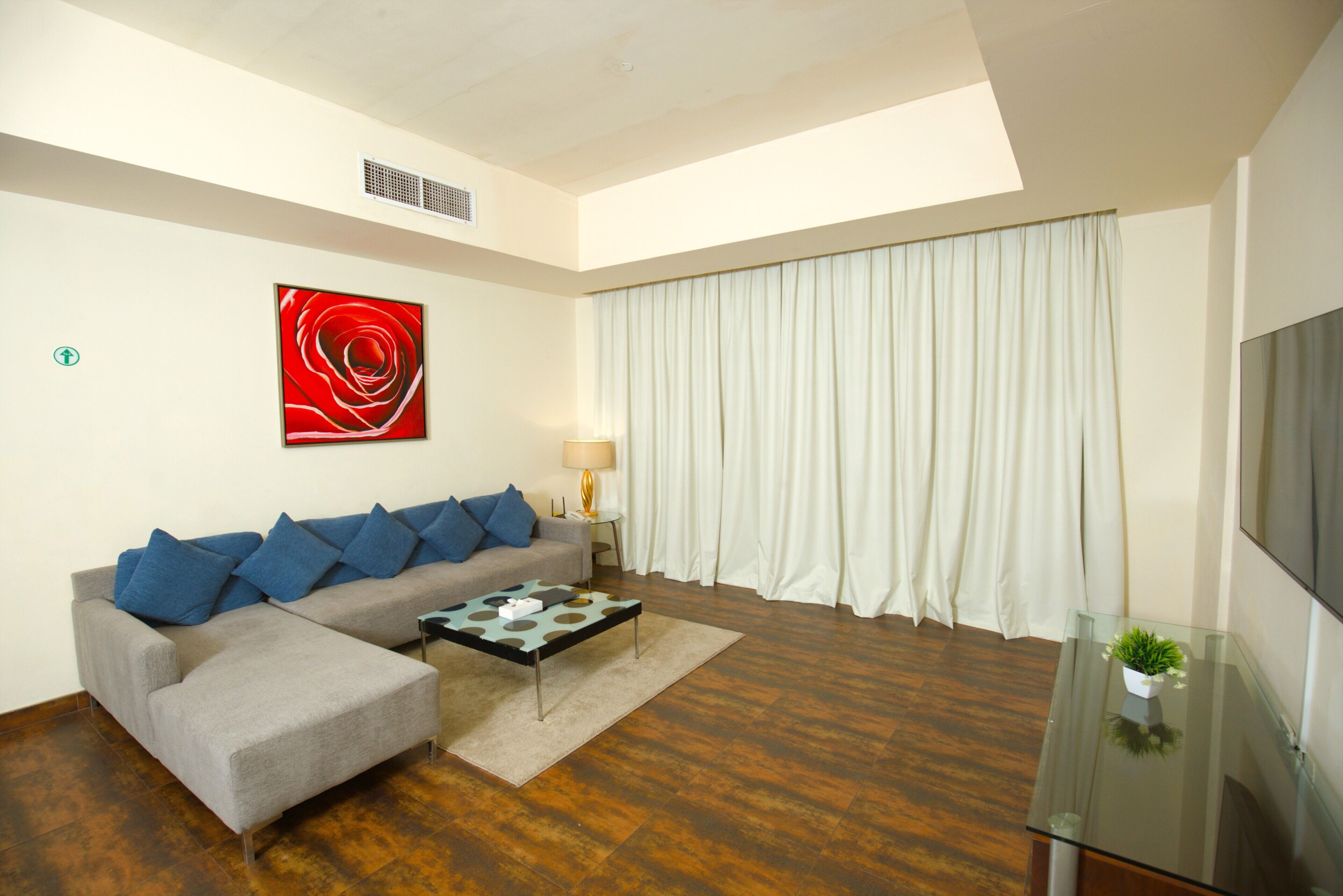 A modern living room with a sectional sofa, a coffee table, a TV on a glass stand, a red abstract painting, and a large window with curtains.