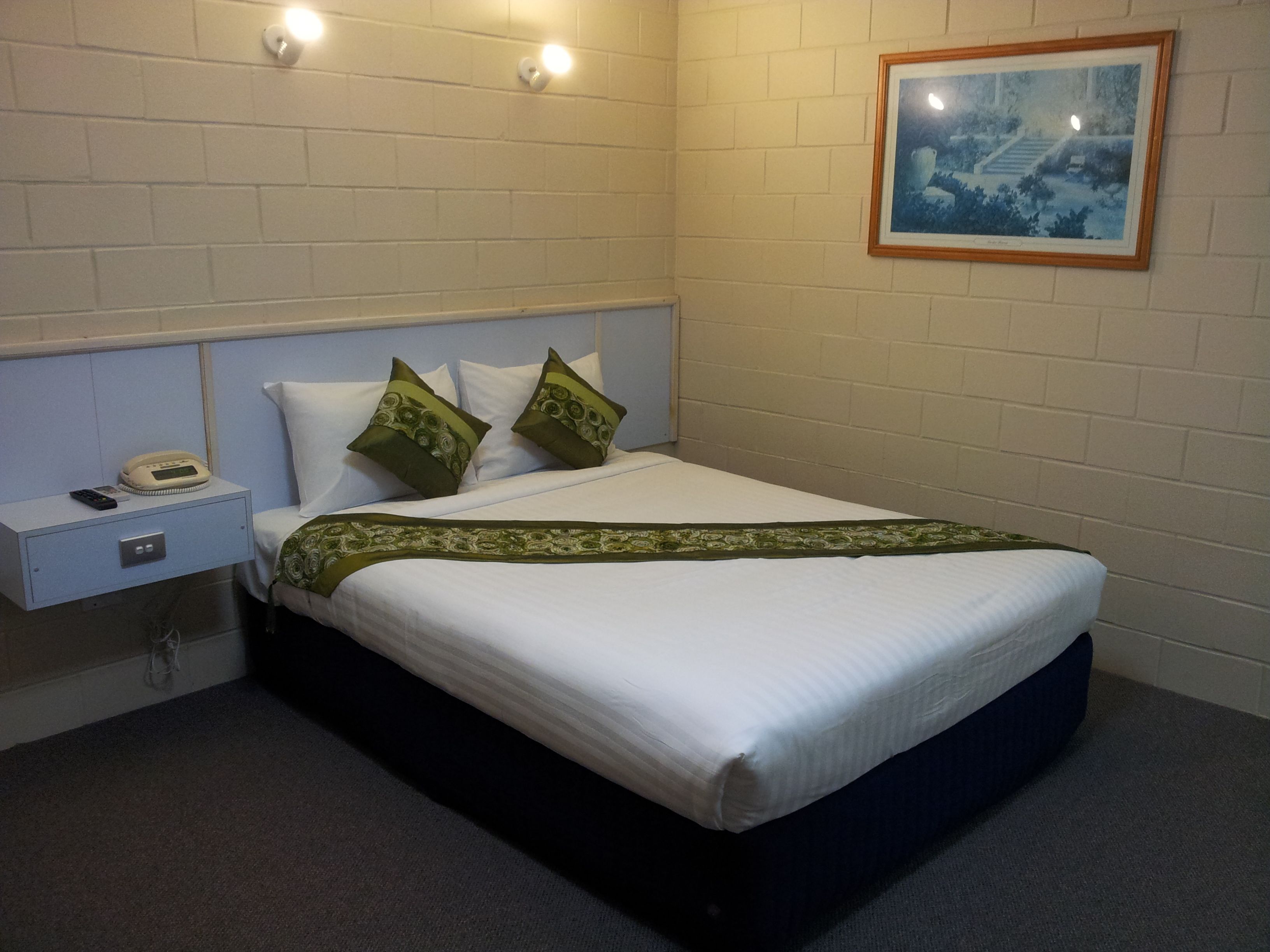 A neatly made bed with white linens and green pillows, a bedside table with a phone, and a framed picture on the wall.