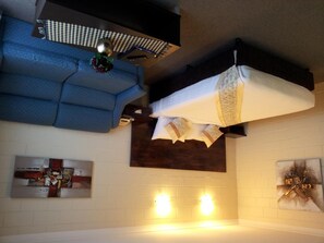 A room with a blue sofa, a white bed with decorative pillows, a wall-mounted shelf with a plant, and a wall art piece.