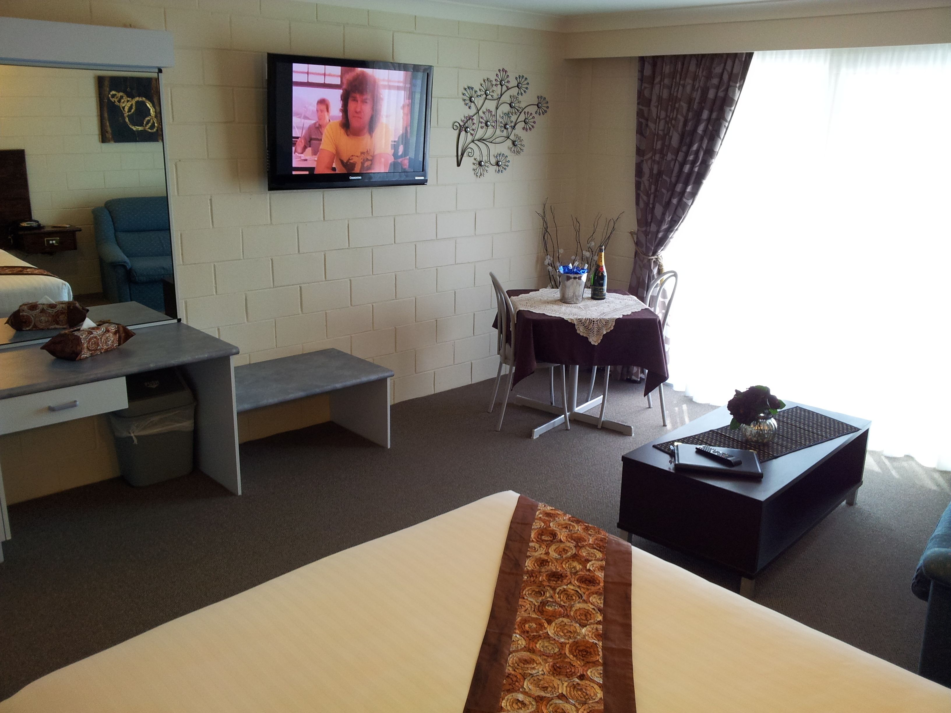A hotel room with a bed, a desk, a chair, a table with a vase, and a television on the wall.
