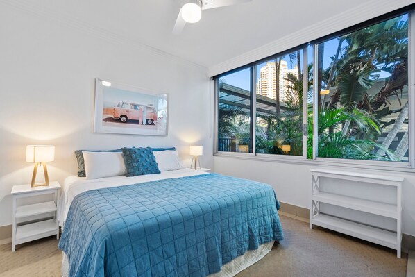 A bedroom with a large window, a bed with a blue quilt, two bedside tables with lamps, and a framed picture on the wall.
