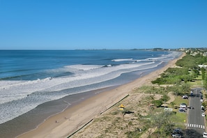 Una carretera costera con una playa a un lado y una calzada bordeada de árboles y edificios al otro.