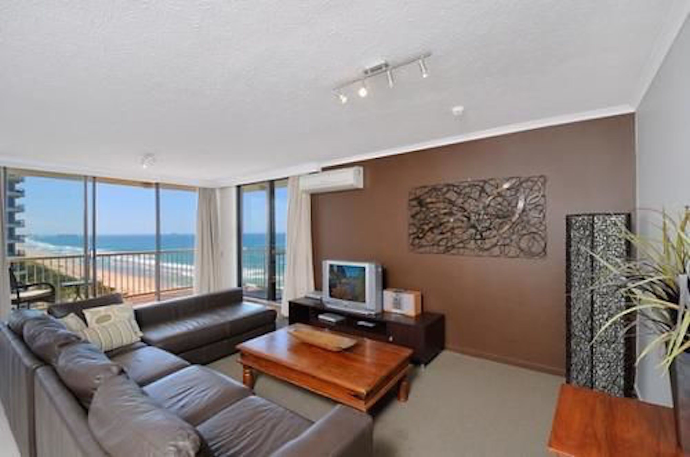 A modern living room with a sofa, coffee table, and a flat-screen TV. There is a large window offering a view of the ocean.
