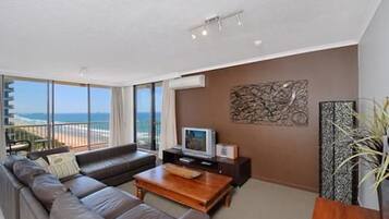 A modern living room with a sofa, coffee table, and a flat-screen TV. There is a large window offering a view of the ocean.