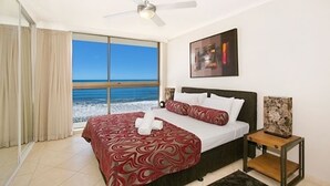 Une chambre d’hôtel avec un lit, un ventilateur de plafond, une fenêtre donnant sur la plage et un tableau encadré au mur.