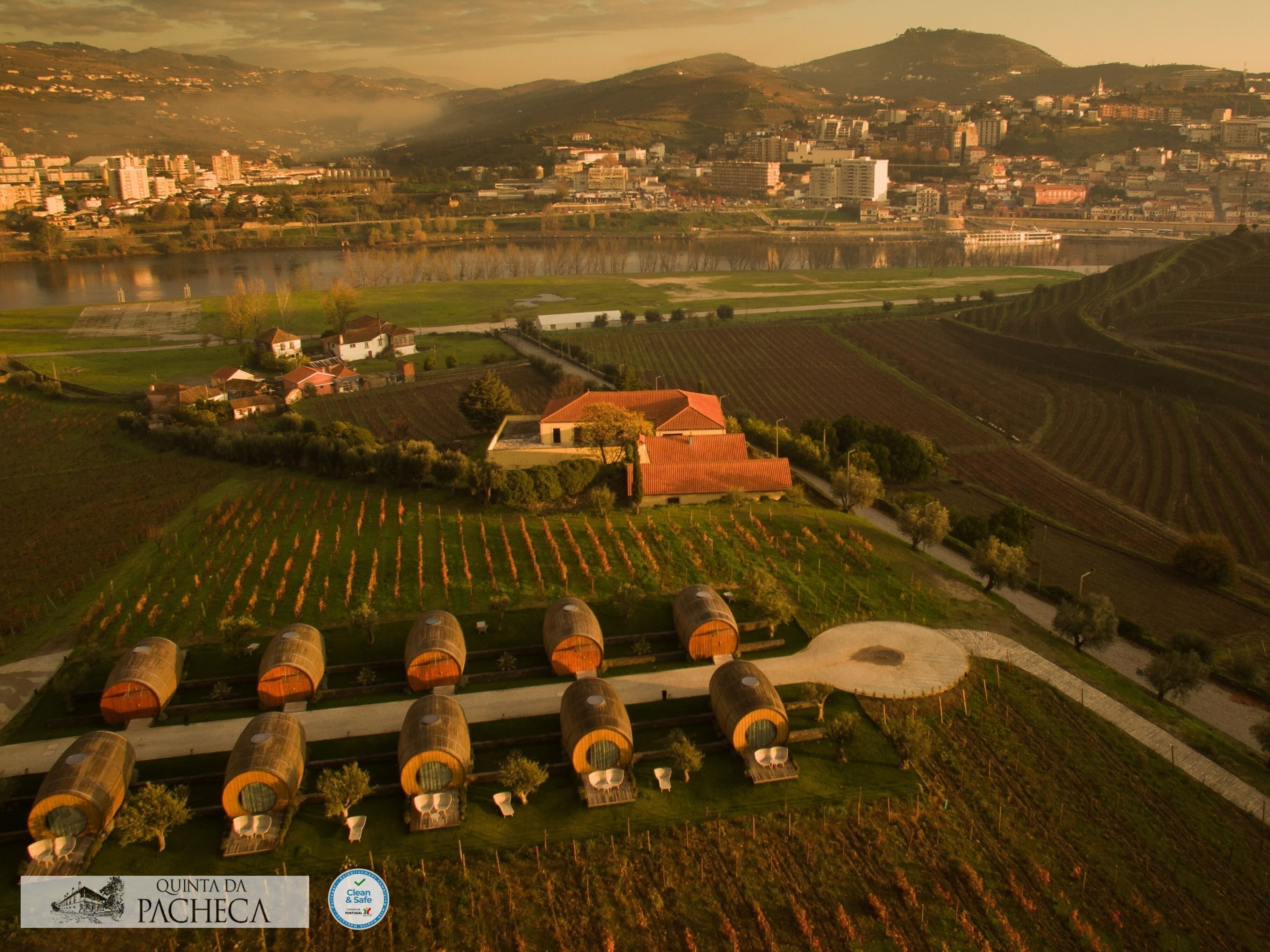 An aerial view of Quinta da Pacheca, featuring terraced vineyards, traditional wine cellars, and a small village with buildings and a river in the background.