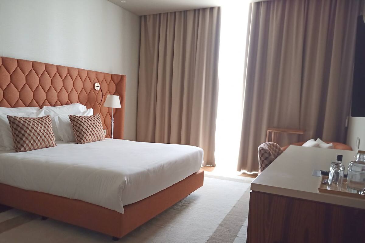 Ein Hotelzimmer mit einem großen Bett, einem Nachttisch, einem Schreibtisch mit Stuhl und einem Vorhangfenster.