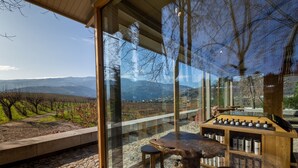 A modern outdoor seating area with a stone floor, wooden table, and stools, overlooking a vineyard and mountains.