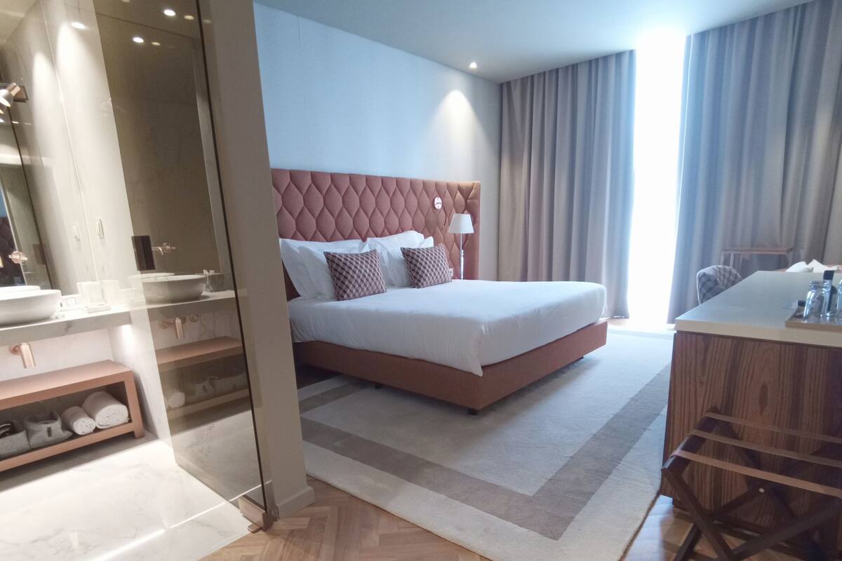 Ein modernes Hotelzimmer mit einem großen Bett, einem Nachttisch, einem Badezimmer mit Waschbecken und Spiegel sowie Blick auf die Stadt.