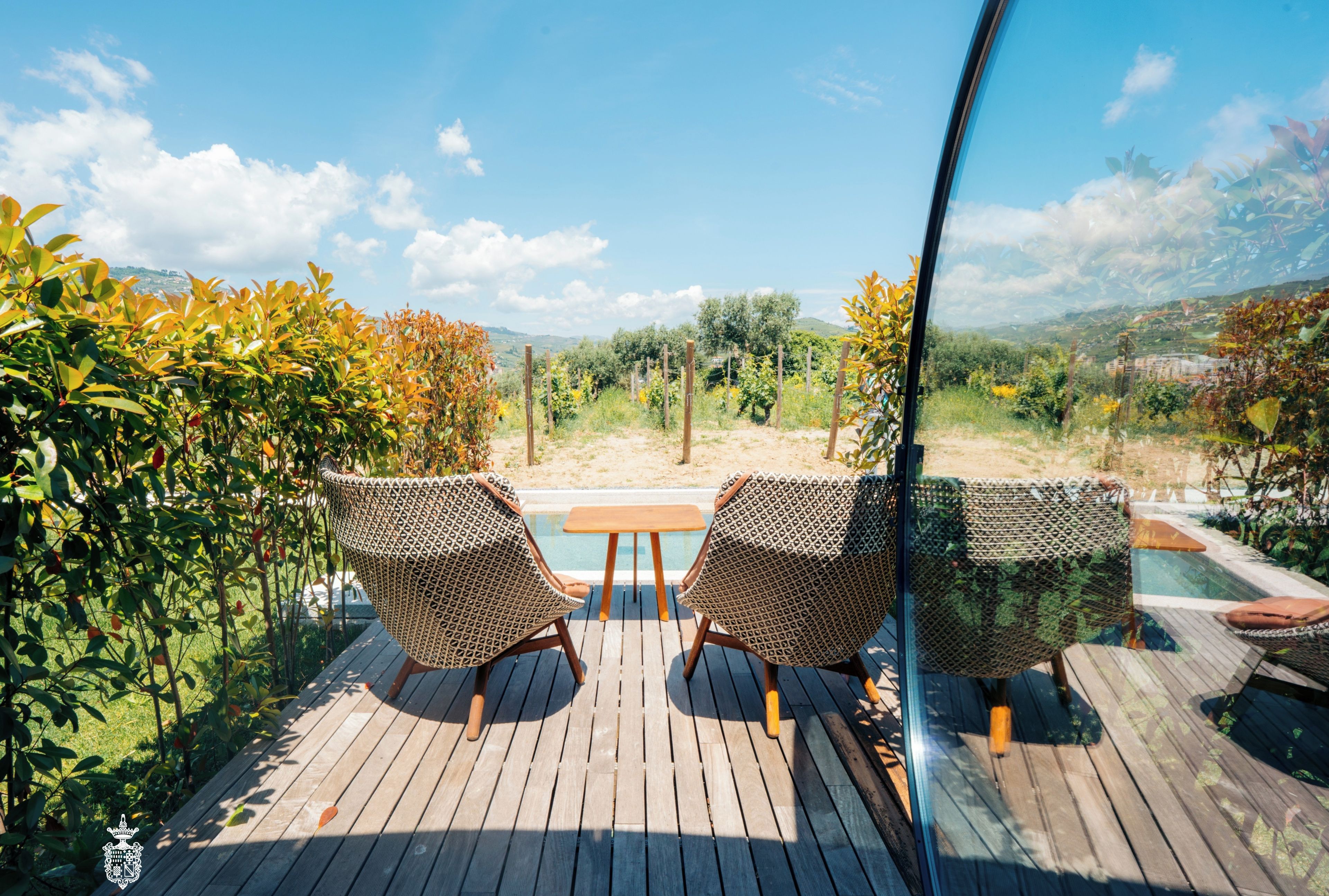 A wooden deck with two wicker chairs and a small table, overlooking a pool and lush greenery.