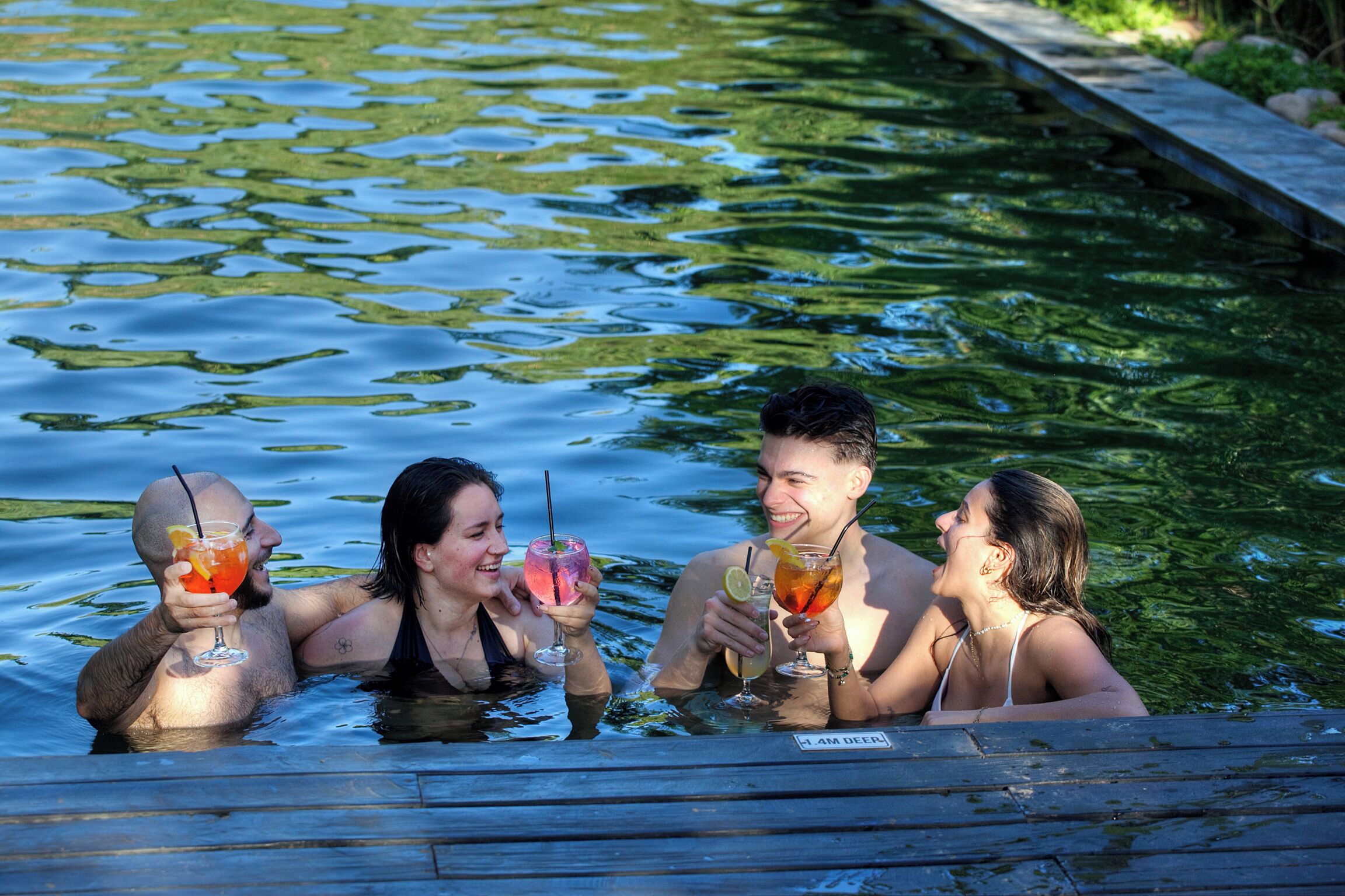 A group of people in a swimming pool with drinks, surrounded by water and a wooden deck.