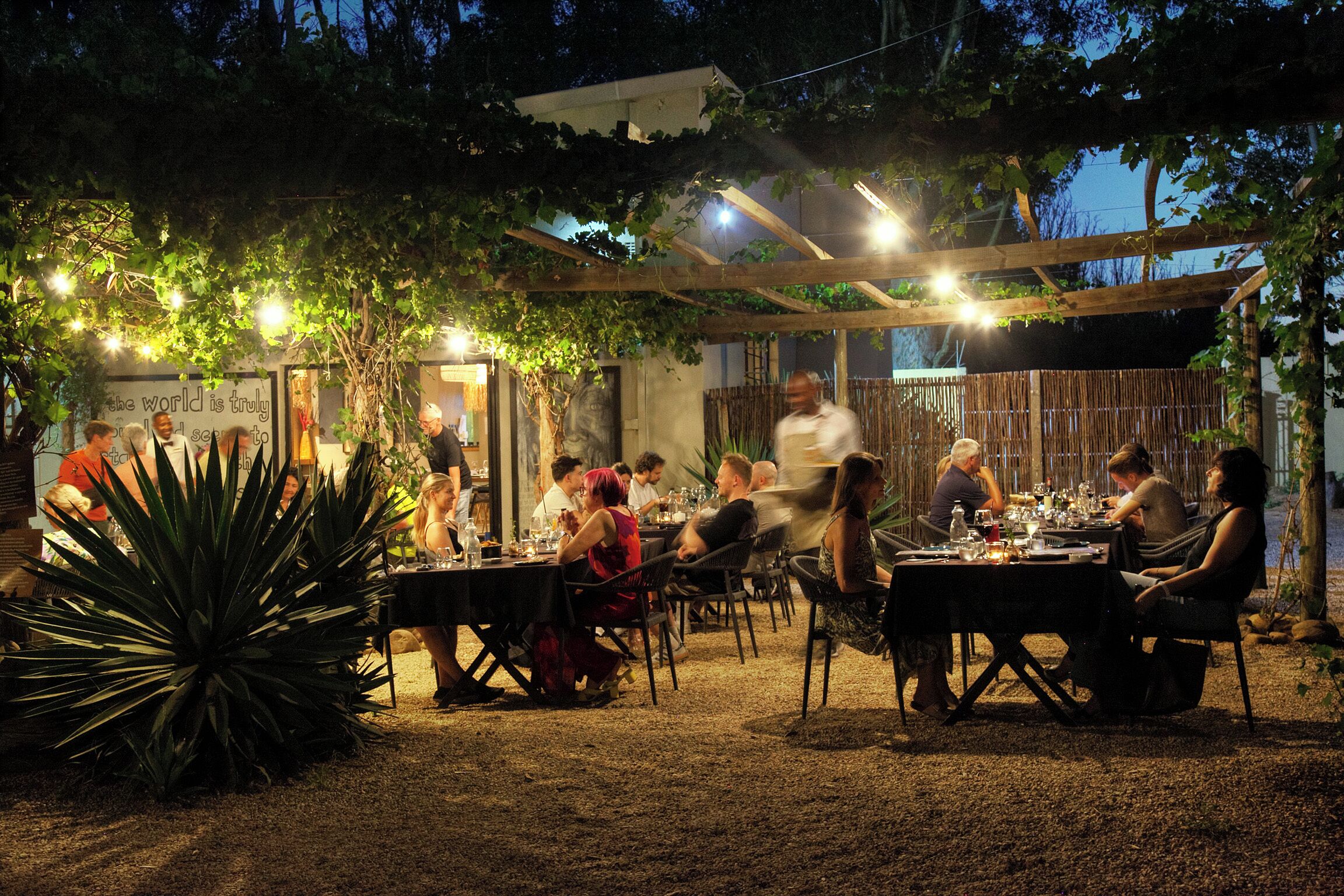 Un espace de restauration en plein air, avec des tables et des chaises, entouré de verdure et agrémenté d’une pergola.