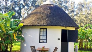 Une maison ronde, coiffée d’un toit de chaume, avec une petite table et deux chaises devant, le tout entouré de verdure.