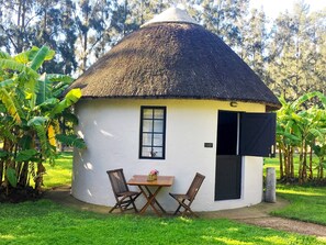 Una casa rodona amb teulada de palla, amb una taula petita i dues cadires al davant, envoltada de verdura.