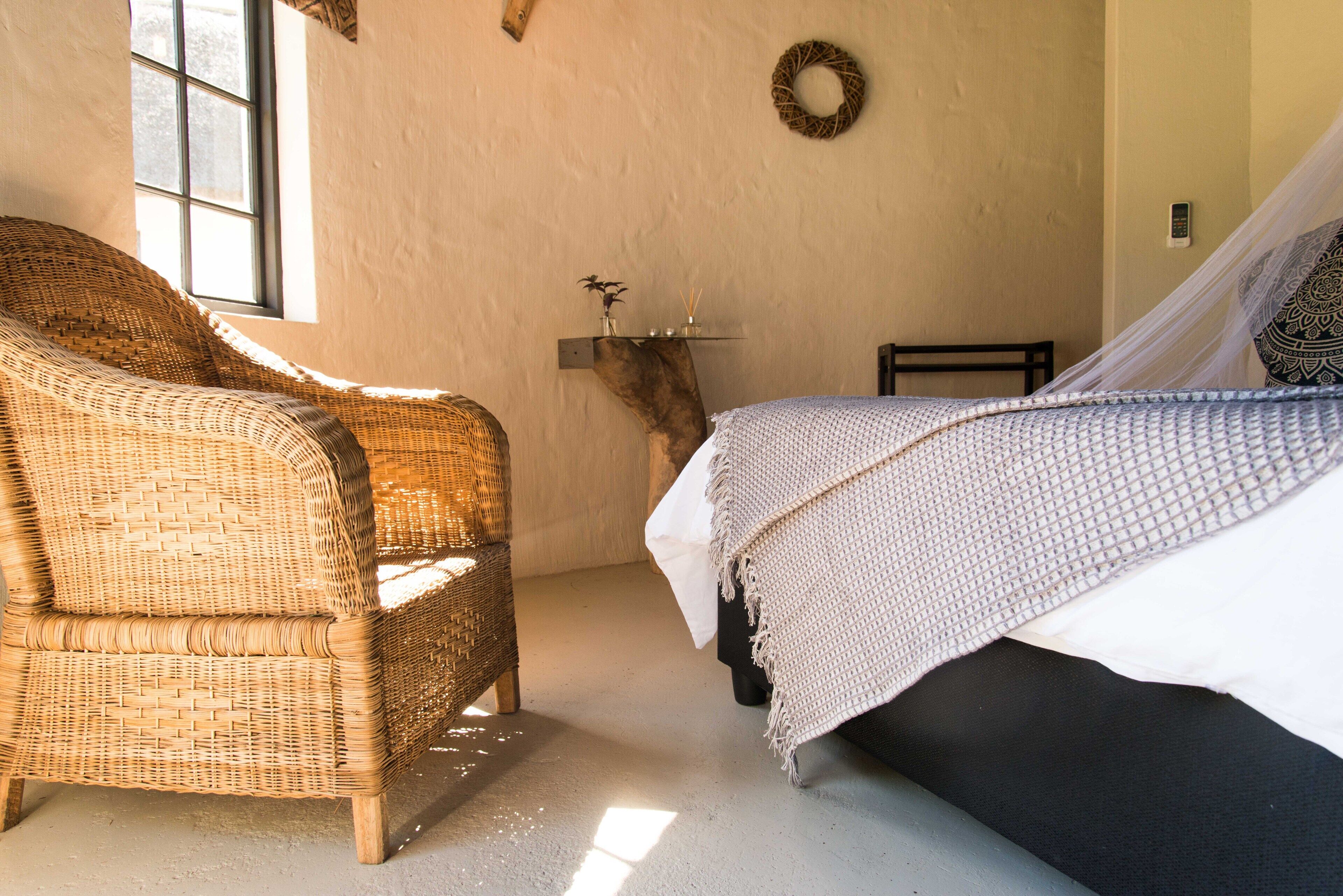 A room with a wicker chair, a bed with a white and patterned bedspread, a wall-mounted shelf with decorative items, and a wall-mounted telephone.