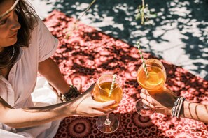 Two people holding glasses of a beverage with a straw, sitting on a patterned blanket by a body of water. - Addo African Home (Addo)