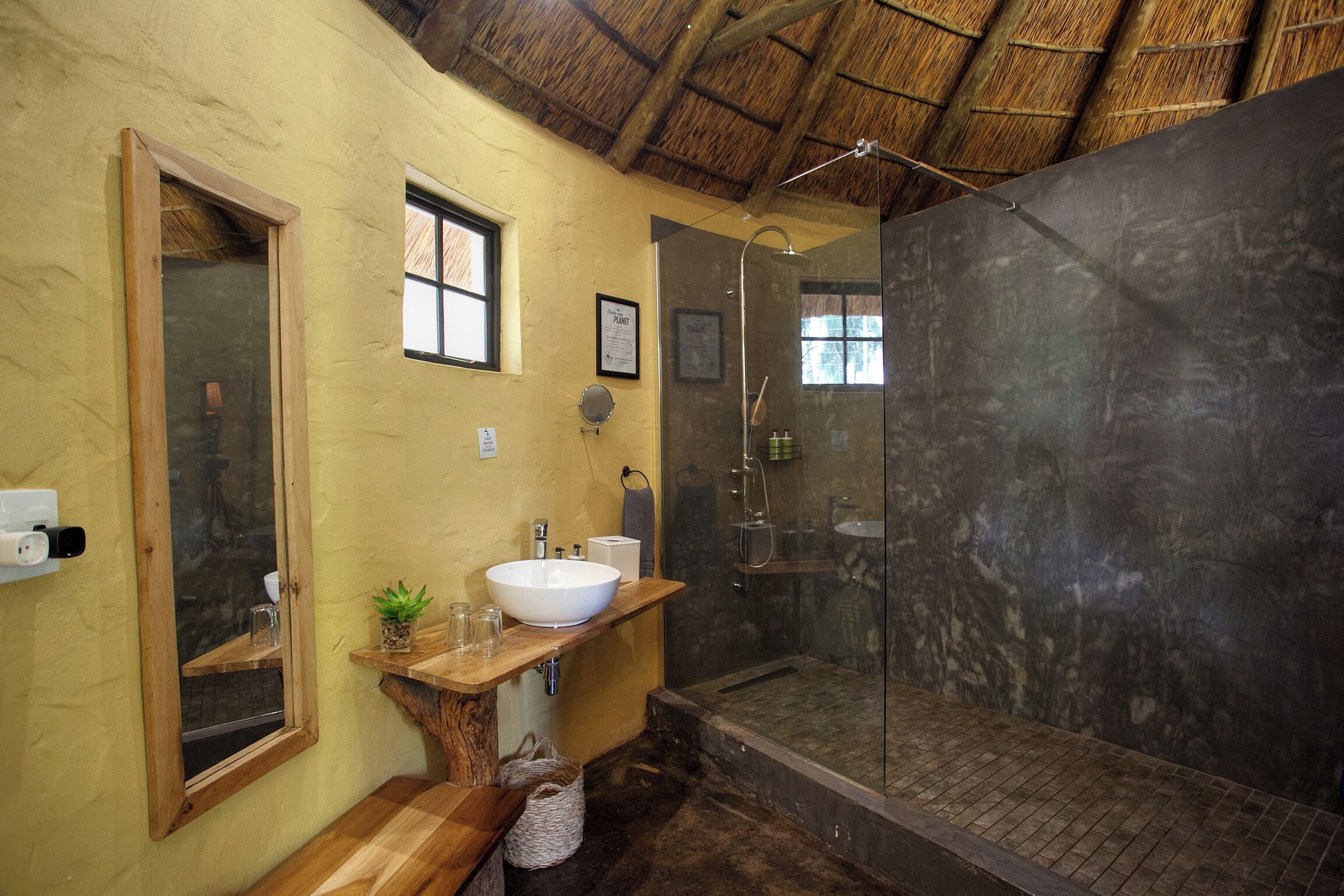 A bathroom with a large mirror, a wooden vanity, a white sink, a shower with a glass door, and a thatched roof ceiling.