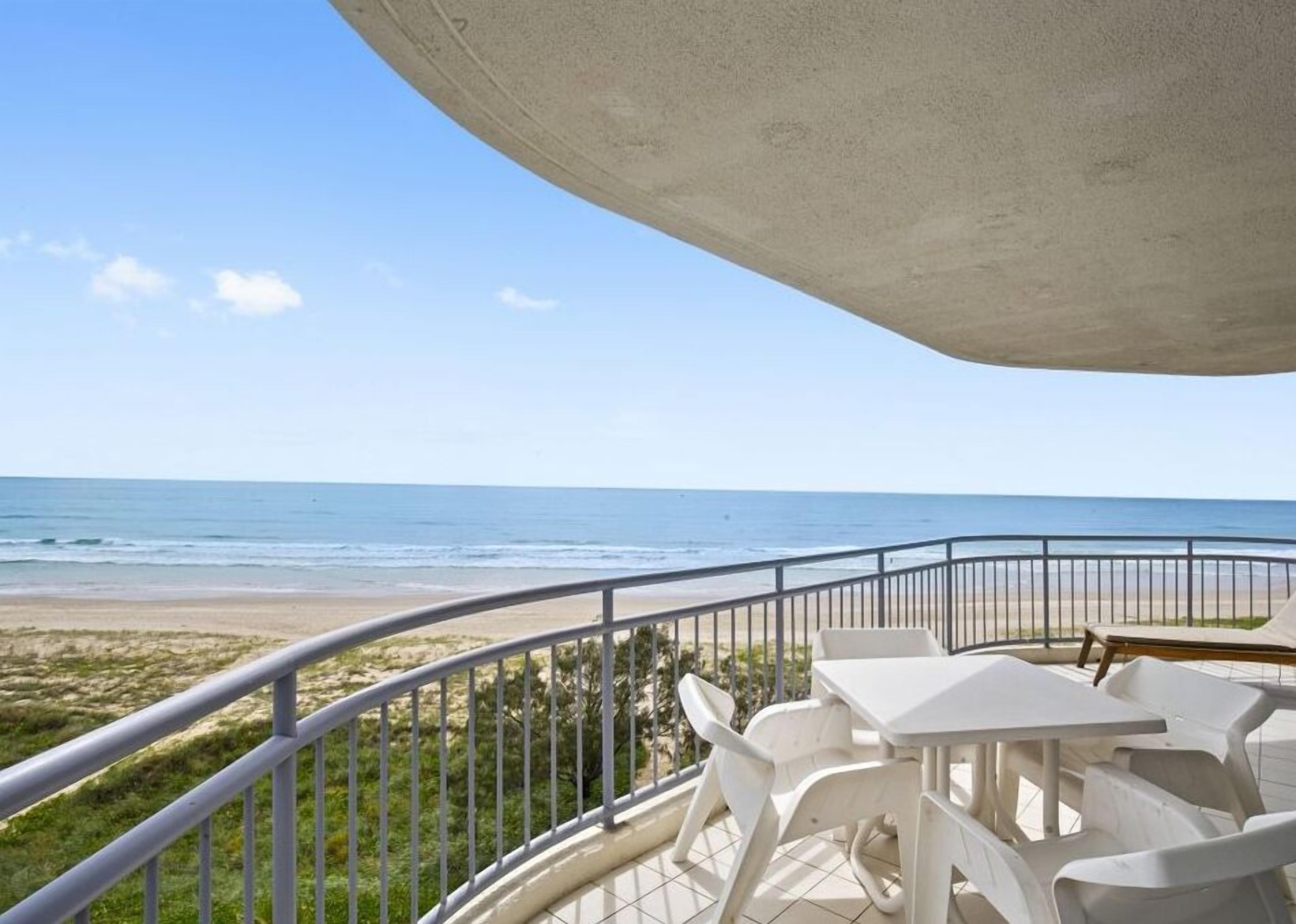 Un balcón con vistas a la playa, sillas de plástico blancas y techo de hormigón.