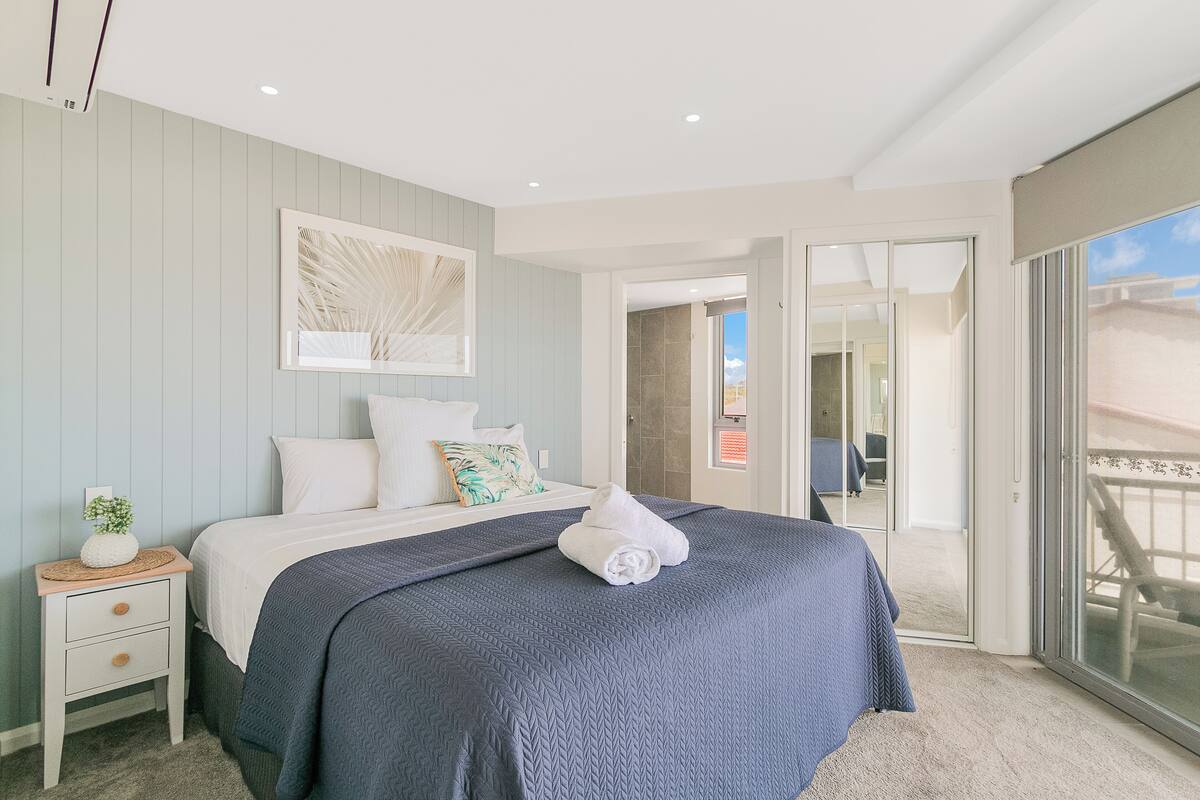 A bedroom with a large bed, a nightstand, a wall-mounted artwork, and a view through a sliding glass door to an outdoor area.