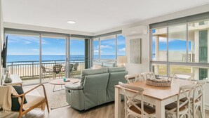 A spacious living area with a large sliding glass door leading to a balcony with a view of the ocean. There is a light green sofa, a wooden dining table with white chairs, and a small round table with a lamp. - Oceanside Resort (Bilinga)