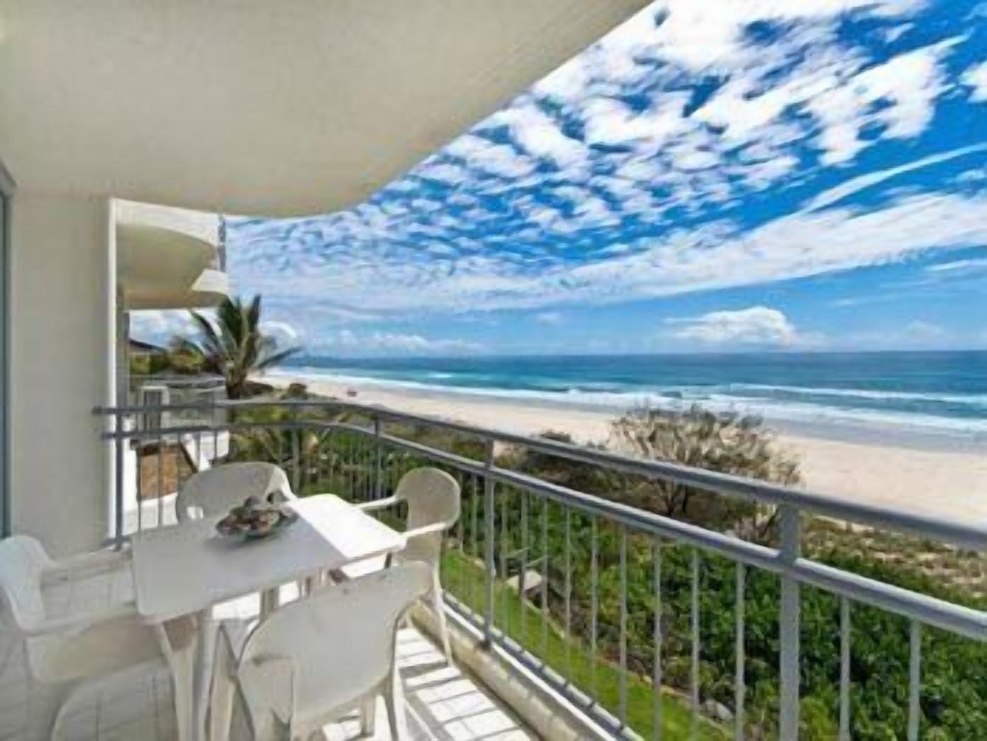 A balcony with a table and chairs, overlooking a beach with waves and a clear sky.
