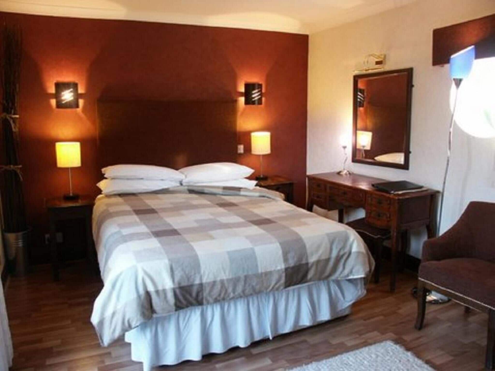 A hotel room with a large bed, a wooden dresser, a desk, and a chair. There are two wall-mounted lamps and a mirror above the dresser.