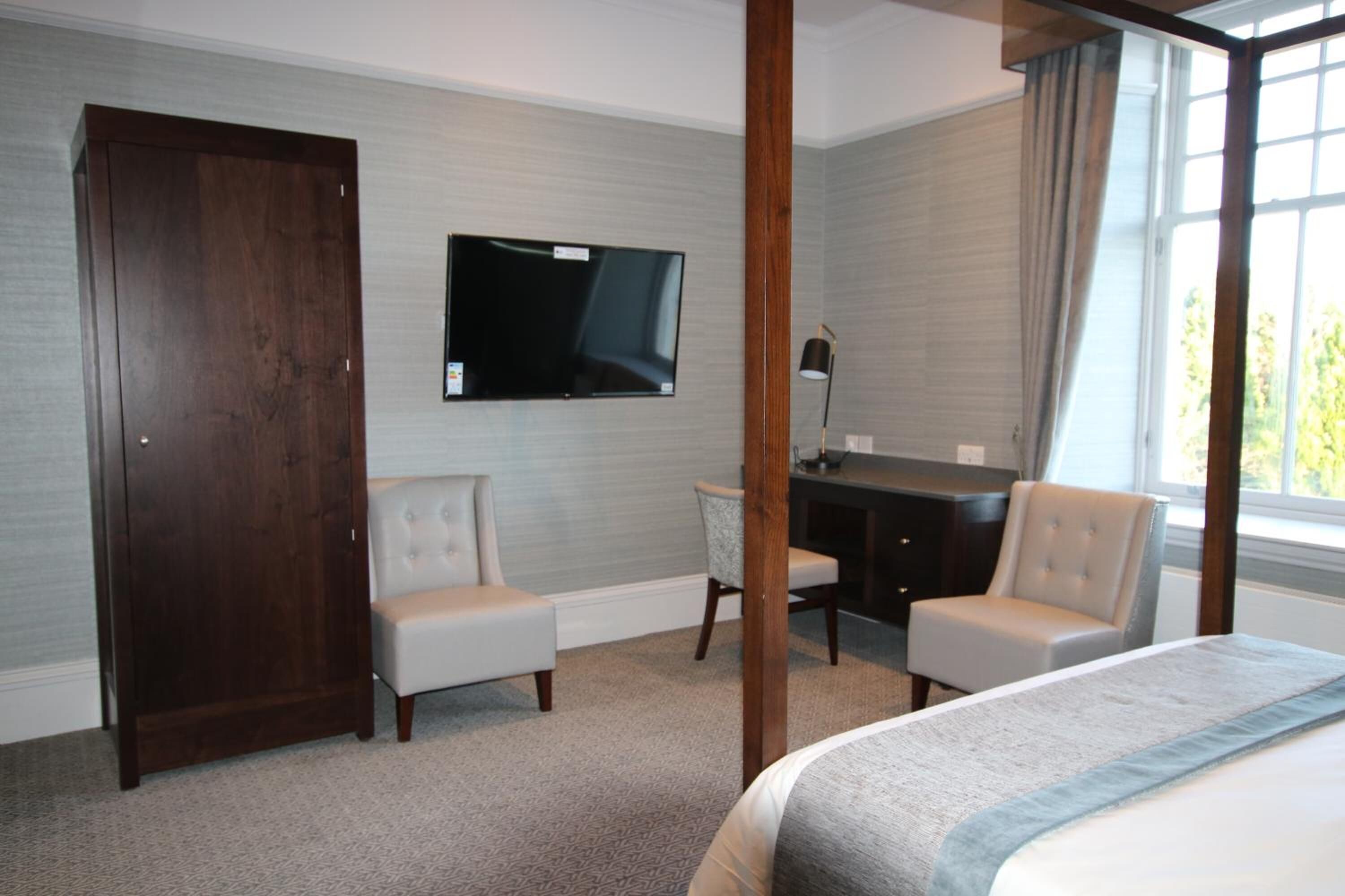 Ein Schlafzimmer mit einem Himmelbett, einem flachbildfernseher an der Wand, einem Schreibtisch mit zwei Stühlen und einem großen Fenster mit Vorhängen.