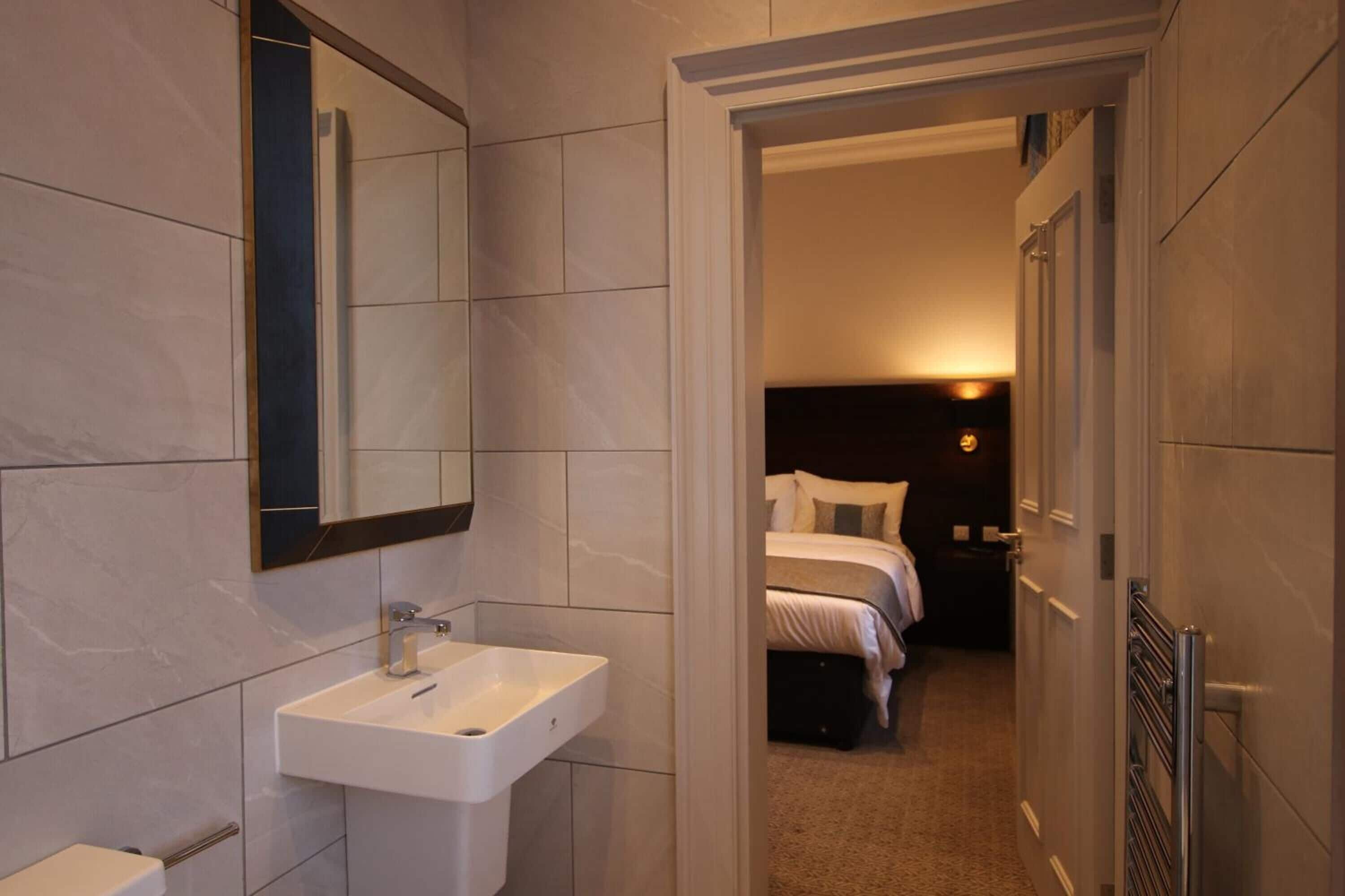 A modern bathroom with a white sink and mirror, a visible toilet, and a hallway leading to a bedroom with a bed and nightstand.